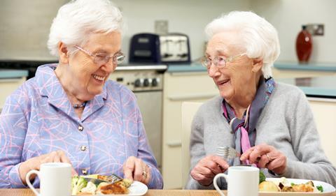 Two older women eating roast dinner with cups of tea, laughing